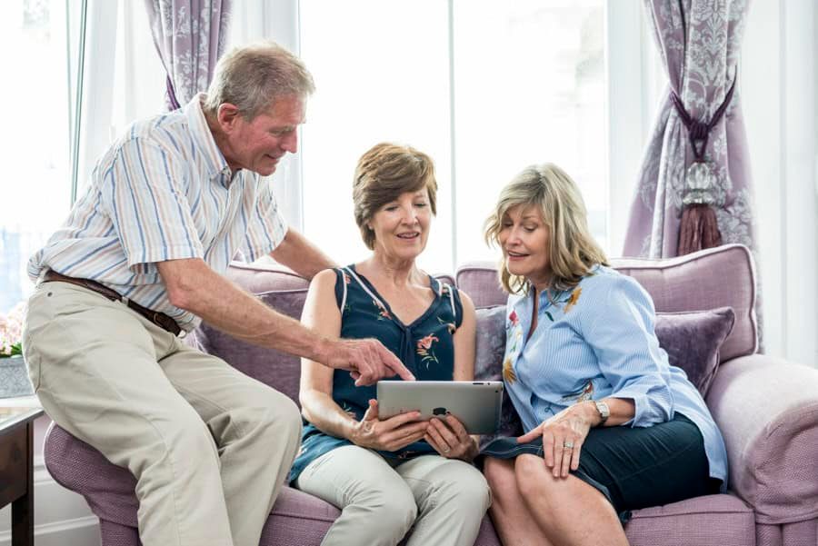 Three elderly people sitting on a purple couch looking at a tablet.