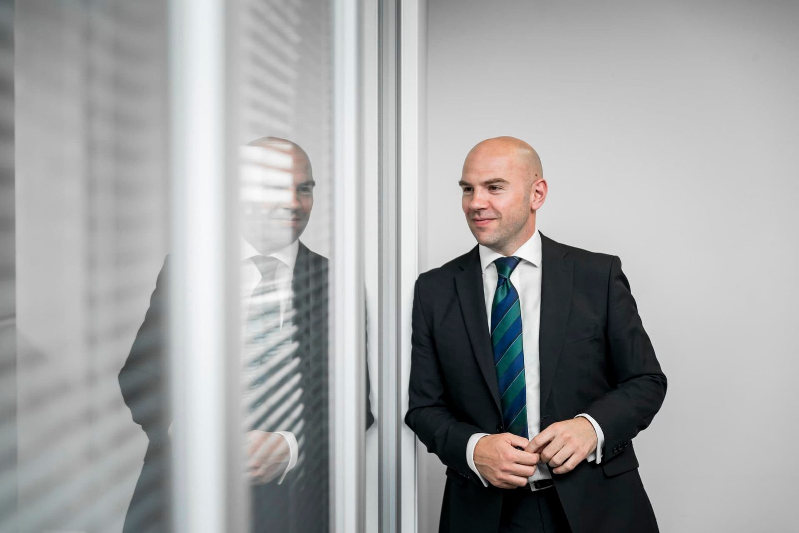 A bald man in a suit standing in front of a window.