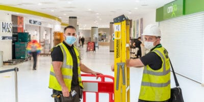 Two men in yellow vests working on a ladder in a shopping mall.
