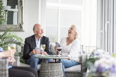 An older couple laughing and drinking wine in a wicker chair.