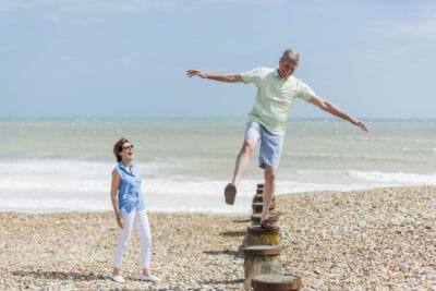 A man and woman jumping on a wooden post on a beach.