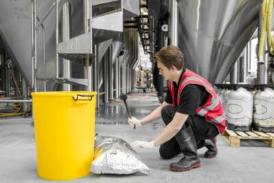 A worker kneeling next to a yellow trash can in a brewery.