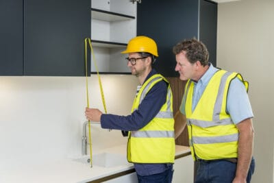 Two construction workers measuring the height of a kitchen cabinet.