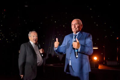 Two men in suits giving thumbs up on stage.
