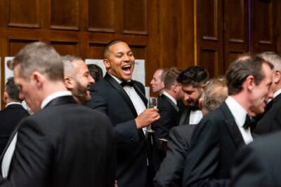A man in a tuxedo laughing in a room full of people.