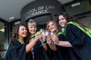 A group of women in graduation gowns toasting champagne in front of the school of law.
