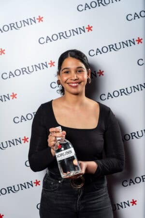 A woman holding a bottle of caorunn in front of a wall.