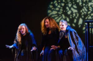 Three women in black cloaks singing on stage.