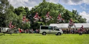 A group of dirt bike riders doing tricks in front of a van.