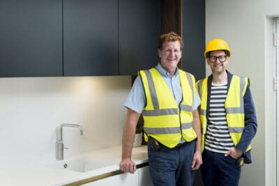 Two men in safety vests standing in front of a kitchen.