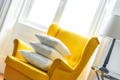 A yellow chair with pillows in front of a window.